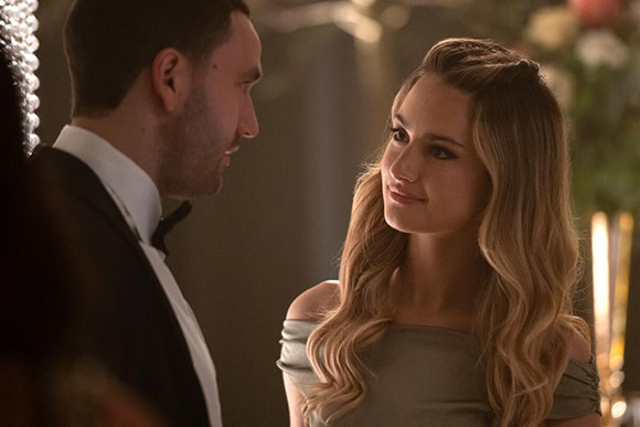 A man in a tuxedo and a woman in a grey dress gaze at one another in a dimly lit setting.
