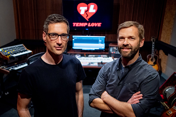 Two men posing in a recording studio with musical equipment and a monitor displaying a broken heart logo.