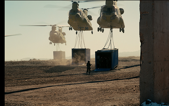 Three large military helicopters hover above a dirt field, lifting blue shipping containers with cables.
