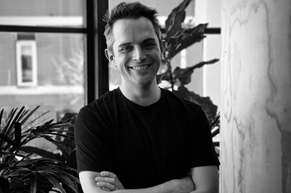 Black and white portrait of a smiling man with short hair wearing a black t-shirt.