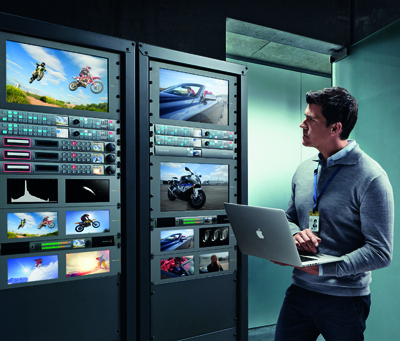 A man holding a laptop while looking at two large server racks displaying various video clips.