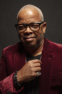 A man with short gray hair and glasses wearing a burgundy velvet blazer and black shirt.