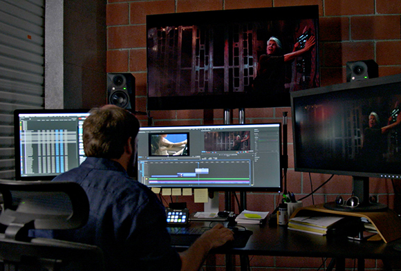A person sits at a desk with three computer monitors displaying video editing software and footage.
