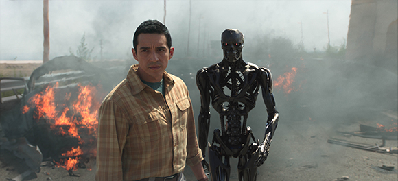 A man in a plaid shirt stands next to a black robotic endoskeleton amidst smoke and fire.