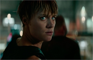 A woman with short blonde hair looks back over her shoulder with a tense expression in a dimly lit setting.