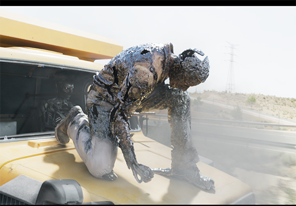 A person coated in thick, dark sludge climbs onto the hood of a yellow truck.