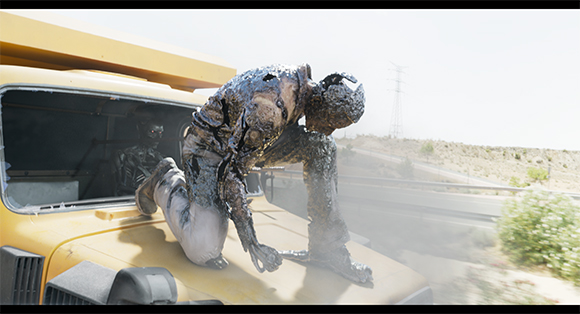 A dark, viscous-looking figure crouches on the hood of a yellow truck near a highway.