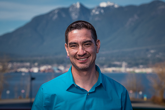 A smiling man in a blue button-down shirt with mountains and water in the background.