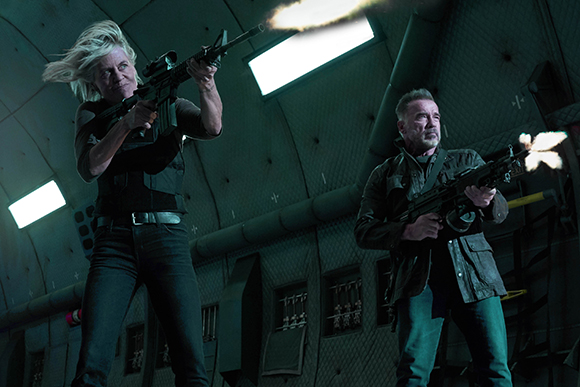 A woman and a man aim and fire rifles in a dimly lit, industrial-looking environment.