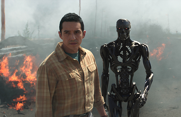 A man in a plaid shirt stands next to a black robotic skeleton amidst smoke and flames.