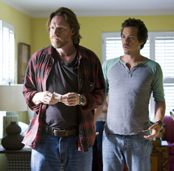 A man in a red plaid shirt and a man in a grey and green shirt stand in a room looking toward the side.