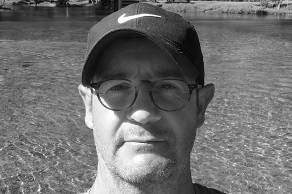 Black and white selfie of a man wearing a Nike cap and glasses with water in the background.