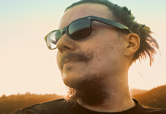 A man with dark hair in a bun and a goatee wearing sunglasses against a sunset sky.