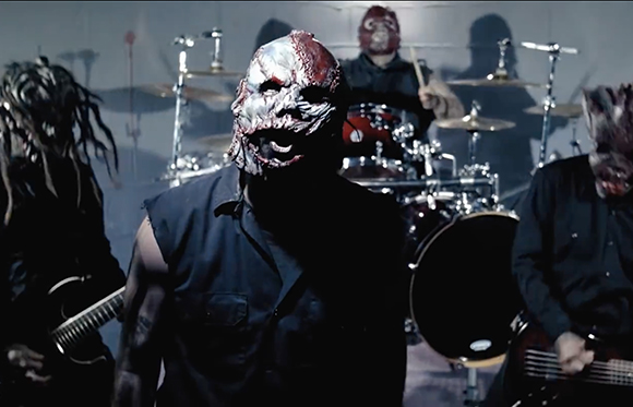 A band wearing grotesque, fleshy masks performs with drums and guitars in a dimly lit studio.