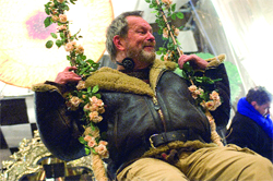 A man wearing a dark shearling jacket sits surrounded by climbing rose vines.