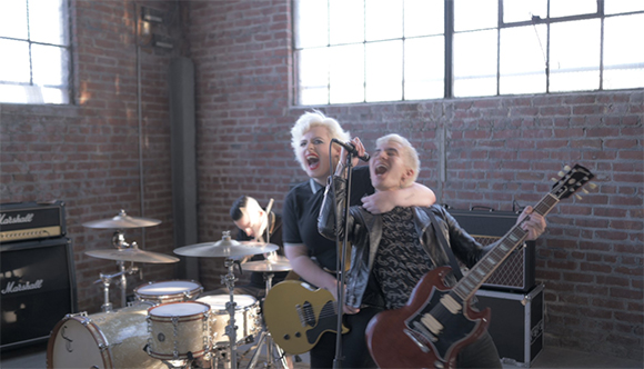 A blonde singer and a guitarist in leather jackets perform in a brick-walled studio with drums and amplifiers.