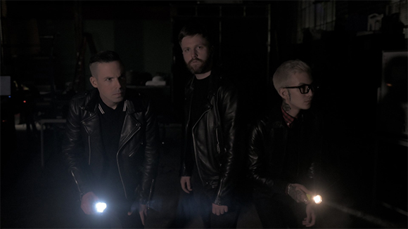 Three men in black leather jackets stand in a dark room, each holding a glowing flashlight.