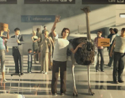 A man in a white shirt waves while standing next to a large ostrich in a busy airport terminal.