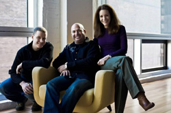 A man and woman sit on a yellow armchair while a young man crouches beside them in a bright room.