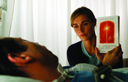 A woman holds a book up for a man lying in a hospital bed with medical tubing.