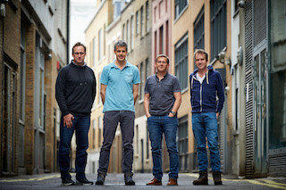 Four men standing in a line in a narrow urban alleyway between tall buildings.