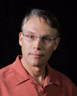 A man with short graying hair and glasses wearing a reddish-orange collared shirt against a black background.