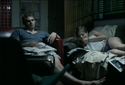A man sits reading a newspaper while a woman lies on a couch in a dark room.