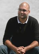 A bald man with glasses wearing a black sweater over a white collared shirt, sitting with hands clasped.