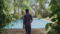A man in a plaid shirt walks toward a bright blue swimming pool in a lush, tropical garden.