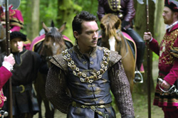 A man in a patterned grey and gold doublet stands in a forest with guards and horses behind him.
