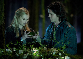A blonde woman and a dark-haired man look down at a small green object in a dark, wooded setting.