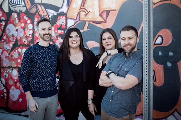 Four smiling adults standing together in front of a vibrant, multicolored graffiti mural.