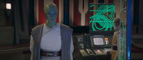 A green-skinned person in white and brown robes stands near a glowing digital screen.