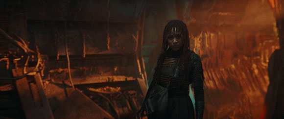 A woman with long braids stands in a dimly lit, metallic, industrial environment.