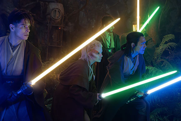 Four people in robes hold glowing yellow, green, and blue lightsabers in a dark forest.