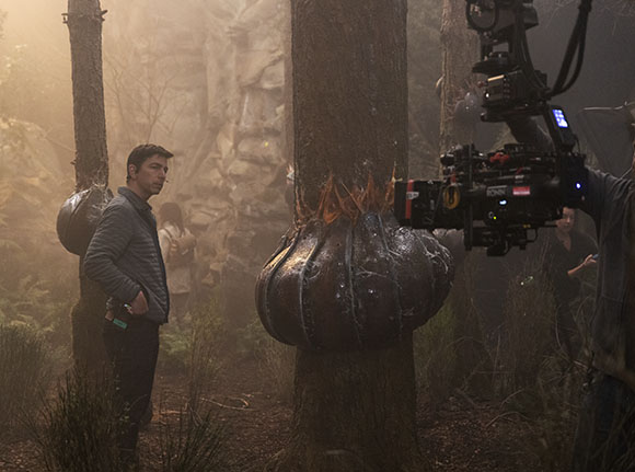 A man in a grey jacket stands in a misty forest next to a large, dark, bulbous object.