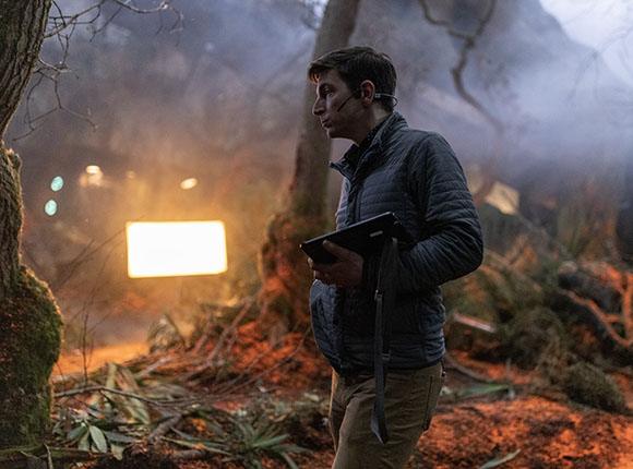 A man in a quilted jacket holds a tablet while standing in a misty, dimly lit forest.