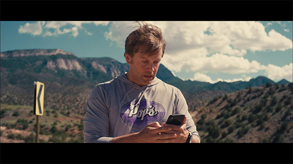 A man in a grey hoodie looks down at a smartphone held in both hands against a mountain backdrop.
