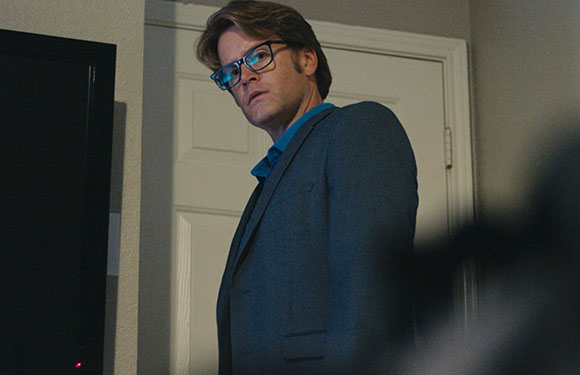 A man wearing glasses and a blue suit jacket looks back over his shoulder in a dimly lit room.