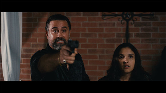 A man points a handgun at the camera while a woman looks on with a startled expression against a brick wall.