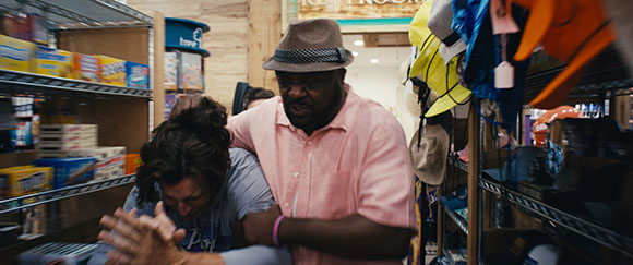 A man in a pink shirt and fedora holds a person in a grey shirt in a retail store.