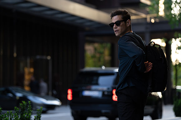 A man in dark clothing and sunglasses looks back over his shoulder while carrying a black backpack.