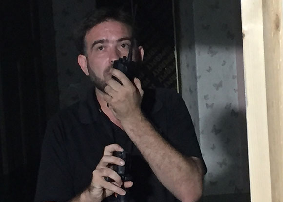 A man in a black shirt holds a walkie-talkie to his mouth in a dimly lit room.