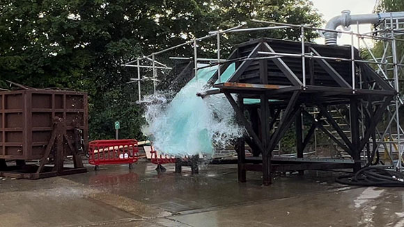 Water erupts from a large black metal industrial structure outdoors near trees.