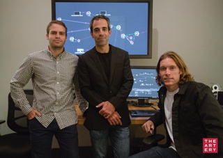 Three men pose in an office with large digital displays showing data visualizations in the background.