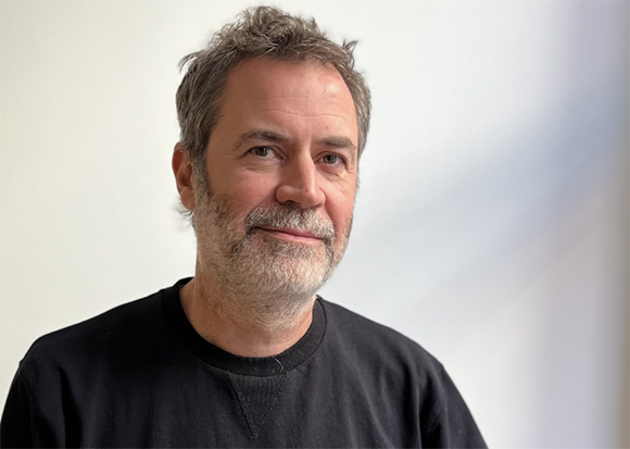 A man with short, salt-and-pepper hair and a beard wearing a black crewneck shirt against a white background.