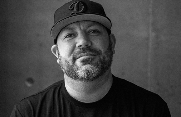 Black and white close-up portrait of a bearded man wearing a baseball cap and a dark t-shirt.