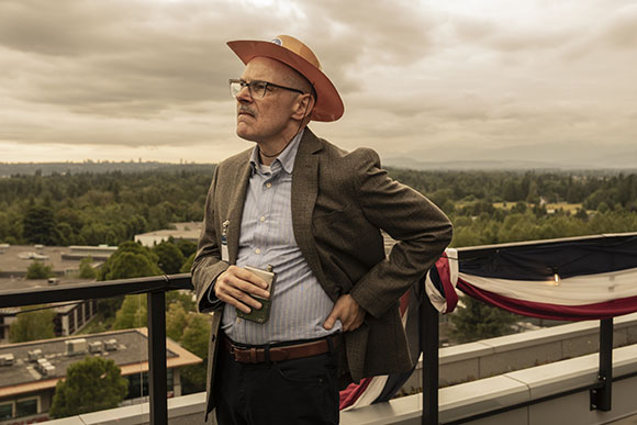 A man wearing a wide-brimmed hat and glasses stands on a balcony overlooking a forest.