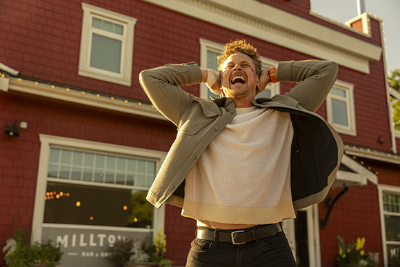 A man laughs joyfully with his hands behind his head in front of a red building.
