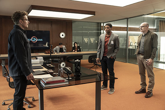 Three men stand in a modern office with wood paneling and a glass partition.